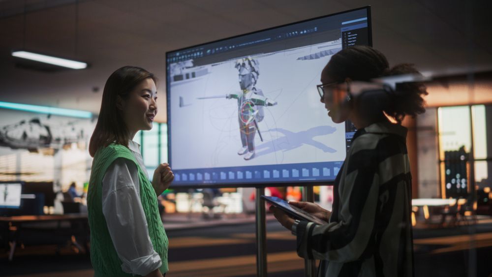 Black Female Game Designer Talking to Asian Female Project Manager And Standing Next To TV Set With 3D Game Character On Screen.