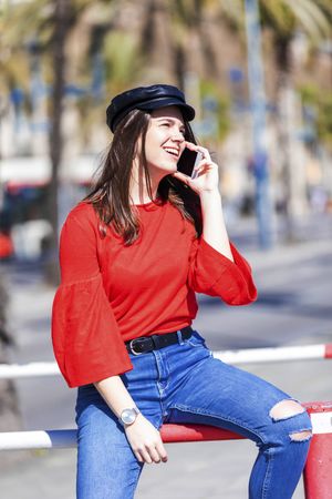 Front view of a beautiful young woman sitting on a metallic fence while using a mobile phone outdoors on a bright day