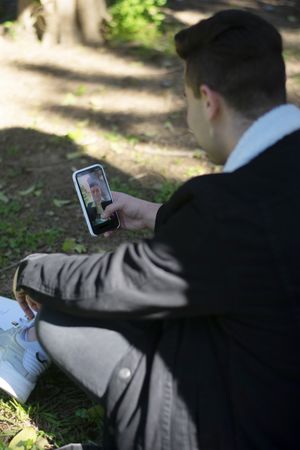 Back view of a young male student sitting on campus while taking video call on a mobile phone