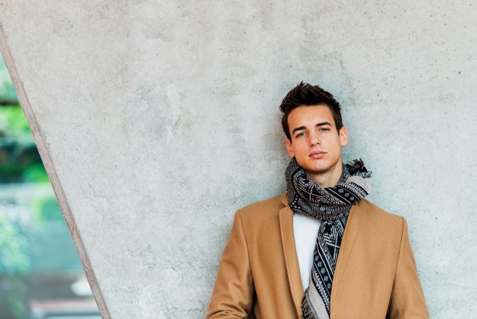 Young man wearing winter coat and scarf looking at camera against cement pillar