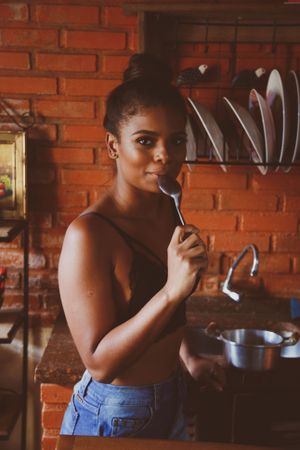 Woman in dark bra and denim pants eating with spoon standing in the kitchen