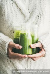 Woman holding three bottles of green smoothie wearing cozy sweater bYvMd0