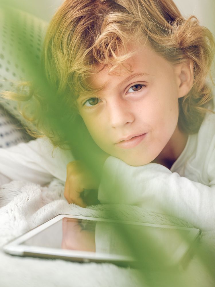 Charming preteen boy lying with tablet and looking at camera