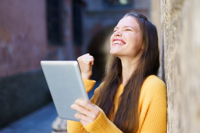 A joyful young woman is celebrating her achievements while happily using a tablet outdoors