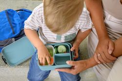 Unrecognizable mother and crop kid sitting on bench with salad lunchbox in daylight
