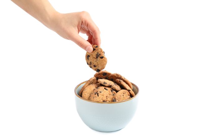 Young woman holding tasty chocolate chip cookie over bowl on plain background, closeup