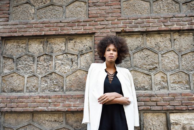 Female in dress and blazer leaning on brick wall and looking in the distance