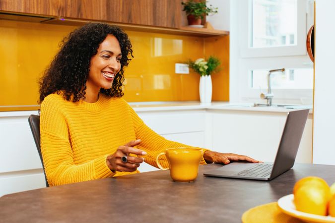 Woman smiling in colorful kitchen sipping from mug in front of her laptop
