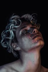 Topless blonde man with UV paint on his face against dark background
