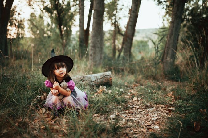 Girl in witch costume crouched down in the forest with her teddy bear