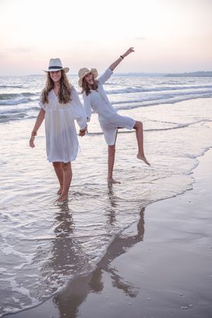 Smiling girlfriends in summer clothes barefoot in water on beach