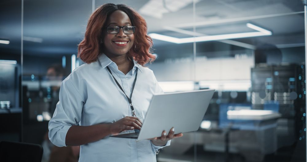 Portrait of a Black Diverse Female Wearing Glasses, Using Laptop Computer, Looking at Camera and Smiling.