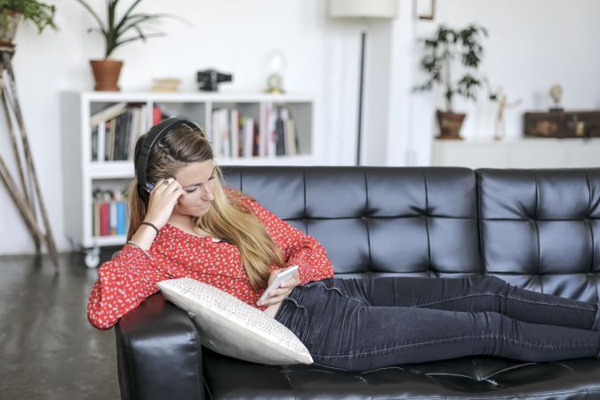 Blonde woman using mobile and listening to music by headphones while lying on leather sofa