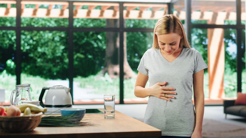 Young Adult Woman with Blond Hair Wearing Gray V-Neck T-Shirt, Rubbing Her Stomach After Food.