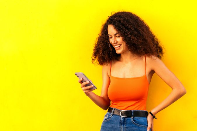 stylish afro latina woman using her mobile leaning on a yellow wall