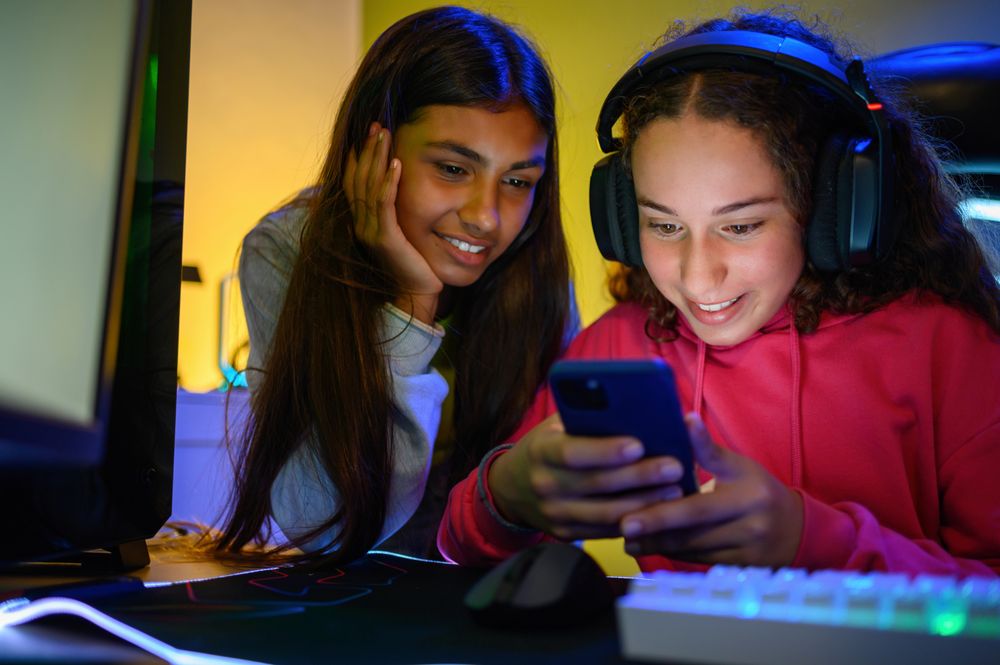 Happy multiracial teenage girlfriends watching smartphone in room with gaming PC
