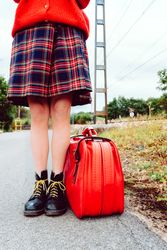 Woman in stylish boots with red suitcase standing on road
