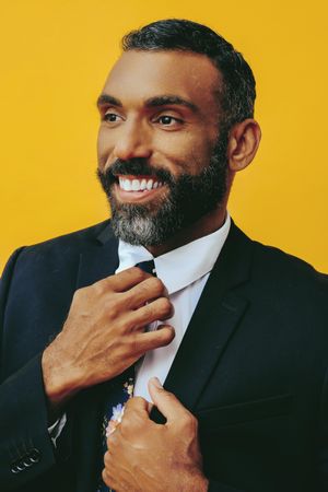 Smiling Black male in suit adjusting his floral tie and looking away in yellow studio