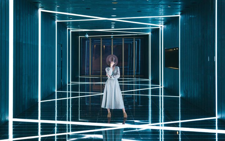 Woman in long dress hiding her face with fedora hat standing in geometrically lit hallway