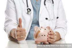Doctor with Thumbs Up Holds Hand to Bandaged Piggy Bank 56GnMe