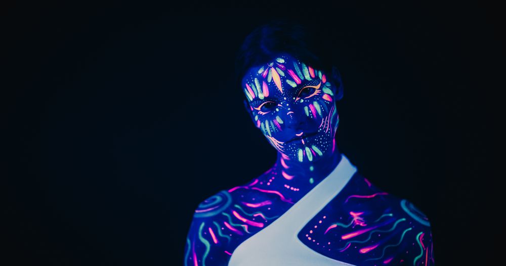 Innovative Beauty Advertising Shoot: Talented Model Showcases Her Unique Style, Neon-Painted Body and Face Contrasting Beautifully with Dark Abstract Backdrop, Creating a Visually Stunning Image