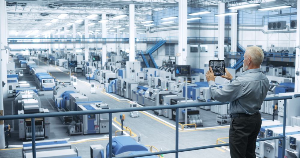 Experienced Male Engineer Standing on a Platform, Using Tablet Computer and Overlooking Production at a Modern Automated Electronics Manufacture with AI Support. Factory Executive at Work