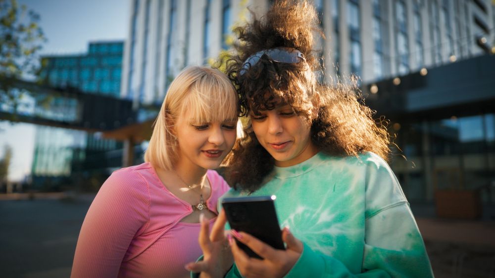 Two Young Women, One With Blonde Hair And The Other With Curly Brown Hair, Are Engrossed In A Smartphone Outdoors