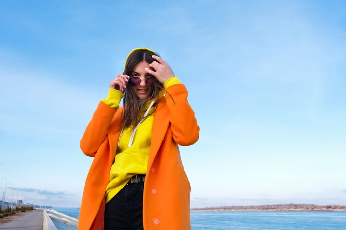 Trendy woman in sunglasses, orange coat, yellow hoodie and jeans against the sea