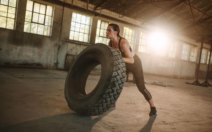 Fit woman doing tire flipping workout at empty warehouse