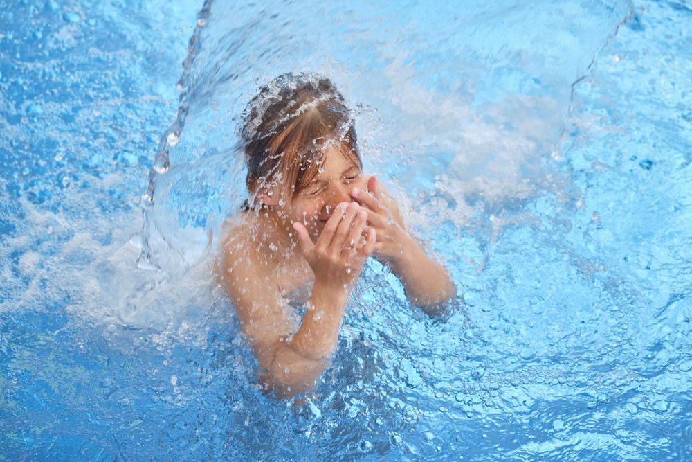 Boy covering nose standing under splashing water in pool
