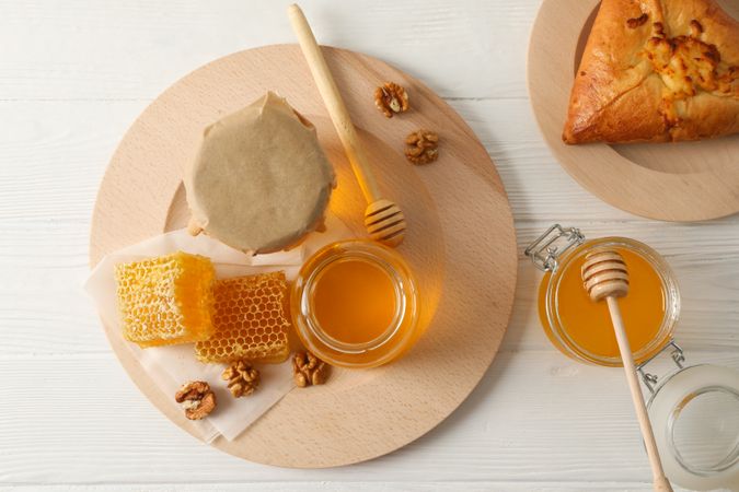 Honeycombs, walnuts, dipper, jars with honey and bun on wooden background, top view