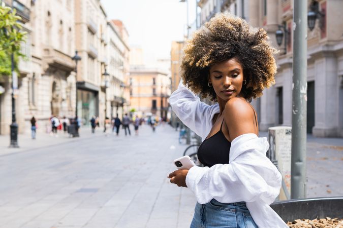 Black woman touching hair while standing on street with smartphone