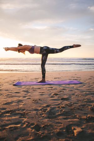 Fitness woman in warrior three pose on yoga mat at the beach