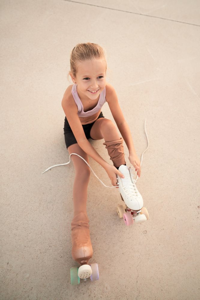 Child lacing up roller skates while sitting on concrete ground
