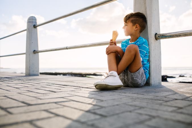 Boy eating an ice cream sitting near a seashore railing with setting sun in the background