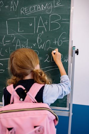 Girl with pink backpack solving math problem on dark board