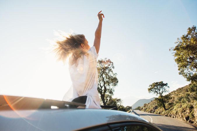 Rearview of woman standing out of sunroof on sunny day with her arms up