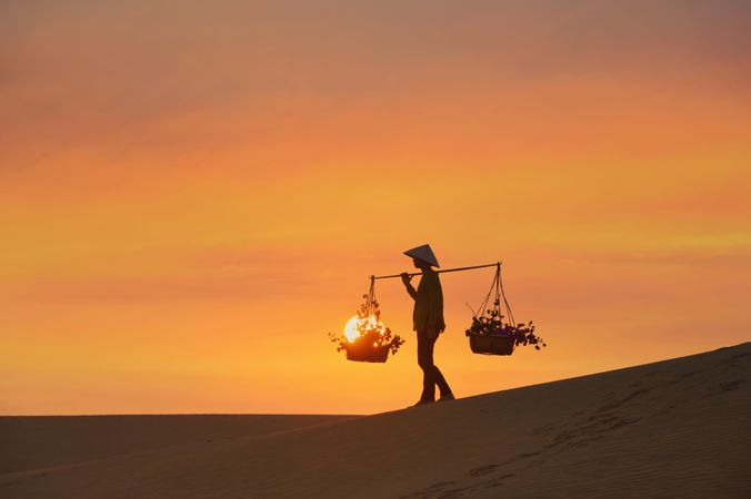 Silhouette of farmer with conical hat holding carrying pole walking on sand dune at sunset