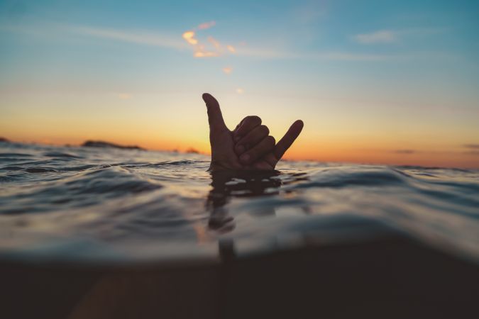 Hand with shaka gesture between water surface and sky at sunset