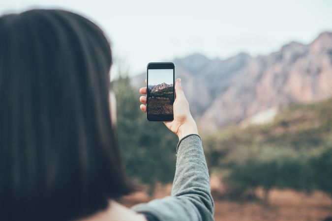 Woman with dark hair taking picture of mountain with smart phone
