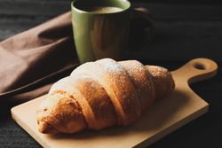 Board with croissant and mug of coffee on dark wooden background, close up