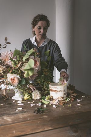 Woman placing plate with cake decorated flower on table with bouquet of blooms in vase