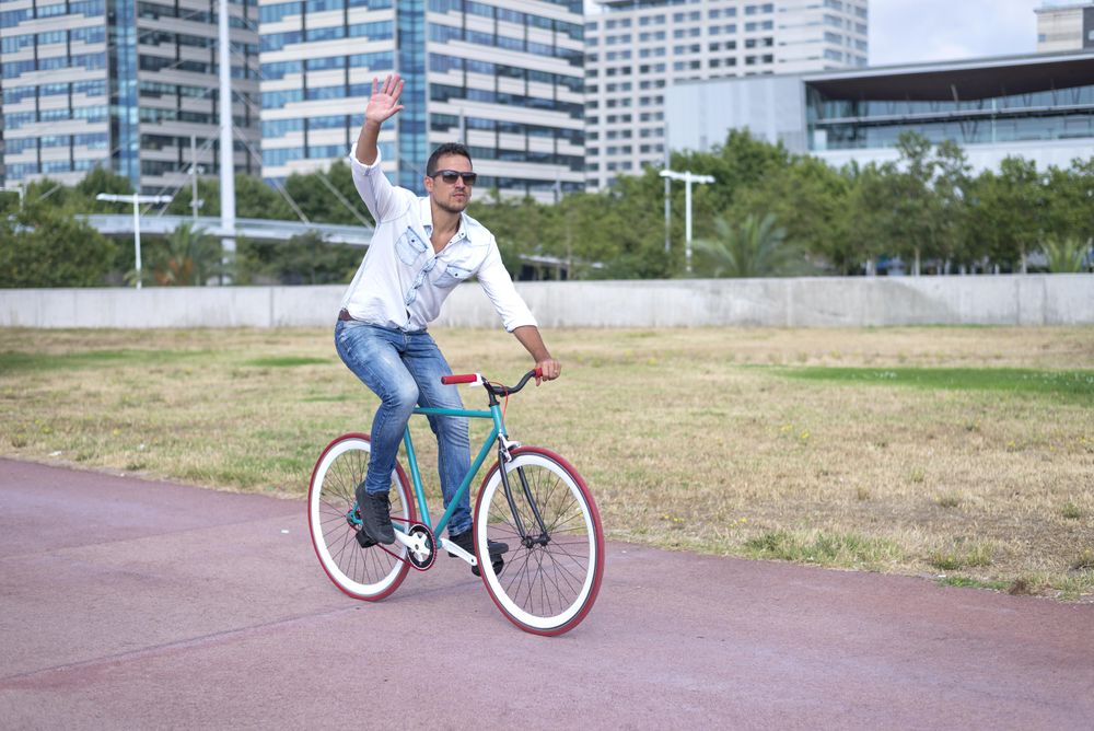 Male riding red and green bike while waving on outside trail next to city buildings
