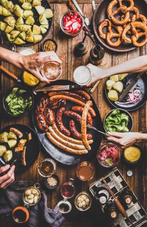 German sausages and pretzels displayed on wooden table with hands holding beer with sausage on fork