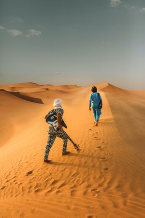 Back of view of person holding a guitar and a person wearing blue outfit walking in desert