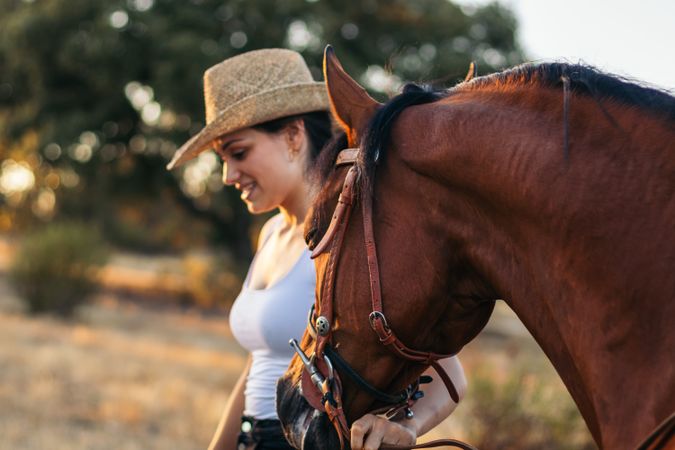 Young woman in hat walking with her horse in the countryside at sunset