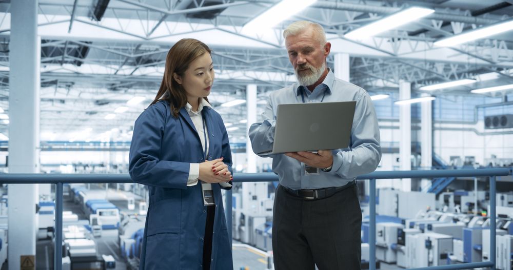 Experienced Male and Female Engineers Standing on a Platform, Using Laptop Computer and Discussing Production at a Modern Automated Electronics Manufacture with AI Support