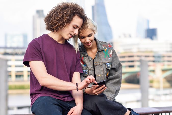Smiling blonde female and curly haired male looking at phone while sitting on fence over river