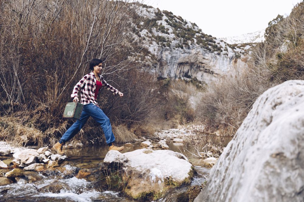 Woman with case jumping on rocks above stream