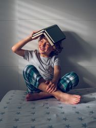 Cheerful boy playing with books sitting on bed in sunlight